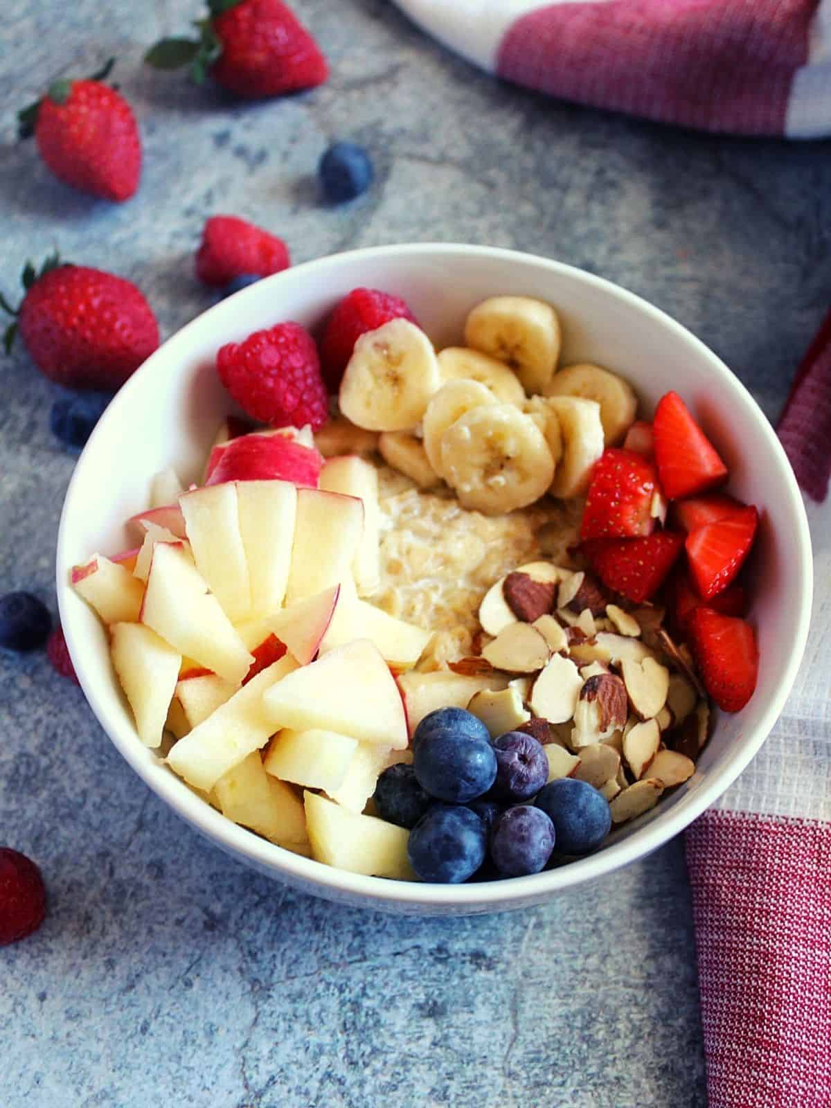 cooked rolled oats served in a bowl with chopped banana, apple, strawberries, blueberries, almonds topping